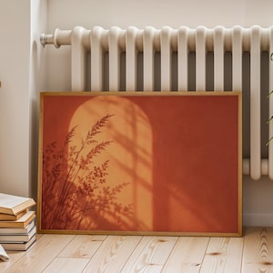 May include: A rectangular framed wall art print featuring a terracotta-colored background with a graphic illustration of an arched doorway and the silhouette of tall grasses casting shadows across the warm, orange-toned surface.