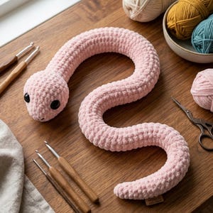 Puede incluir: Un juguete de serpiente de ganchillo rosa claro hecho a mano con ojos de seguridad negros. La serpiente de peluche se exhibe sobre una superficie de madera con ganchos de ganchillo, hilo y tijeras. La serpiente mide aproximadamente 76 cm de largo.