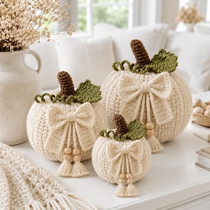 May include: Three crocheted pumpkins in varying sizes, each featuring a cream-colored body, a brown stem, a green leaf, and a curly vine. Each pumpkin is decorated with a cream-colored crocheted bow, wooden beads, and a small tassel hanging down.