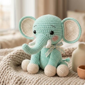 Op de afbeelding: Een gehaakt olifantenspeelgoed in tinten van teal en crème. De olifant heeft grote, afgeronde oren, een slurf en kleine slagtanden. Het heeft zwarte veiligheidsoogjes, roze wangen en een crèmekleurige strik. De olifant zit op een getextureerde, crèmekleurige deken.