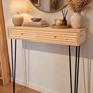 May include: A light-colored wooden console table featuring a horizontal ribbed drawer front with two small round knobs, supported by four slender black metal hairpin legs. The table surface is rectangular with a natural wood grain finish.