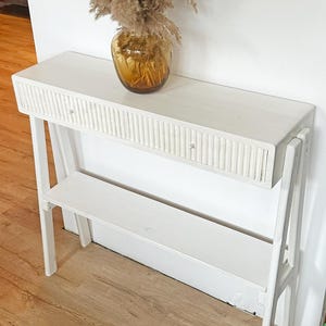 Op de afbeelding: Een witte sidetable met één lade voorzien van een verticaal geribbelde voorkant en een kleine ronde knop. De tafel heeft een onderste plank en schuine poten. Een amberkleurige glazen vaas gevuld met gedroogd pampasgras staat op het blad.