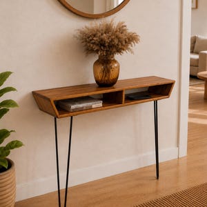 Puede incluir: Una mesa consola de madera con una superficie rectangular y dos compartimentos de almacenamiento abiertos, sostenida por cuatro patas delgadas de metal negro tipo horquilla. Un jarrón de vidrio marrón con hierba de la pampa seca está encima.