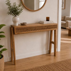 Puede incluir: Una mesa consola de madera de color marrón claro con un cajón único de textura vertical acanalada. La mesa se apoya sobre cuatro patas inclinadas y sostiene un jarrón blanco con vegetación, un cuenco pequeño y dos libros con una vela.