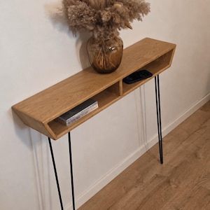 May include: A narrow wooden console table with a light brown wood grain finish and two open storage compartments. The table is supported by four thin black metal hairpin legs. A glass vase with dried pampas grass sits on the top surface.