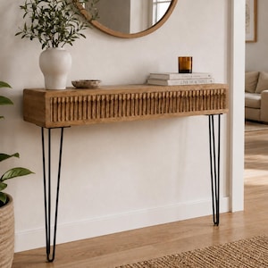 May include: A rectangular wooden console table featuring a textured, vertically grooved front panel and four slender black metal hairpin legs. The table surface holds a white ceramic vase with greenery, a small bowl, and two stacked books.