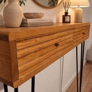Könnte beinhalten: Ein Holzkonsolentisch mit einer texturierten, horizontal gerippten Frontplatte und zwei kleinen runden Knöpfen. Der Tisch steht auf vier schlanken Haarnadelbeinen aus schwarzem Metall. Obenauf stehen eine beige Keramikvase, eine Steinschale und zwei Bücher.