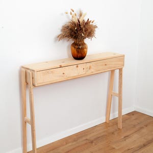 Könnte beinhalten: Ein heller Holzkonsolentisch mit zwei Schubladen, die eine strukturierte Vorderseite aufweisen. Der Tisch steht auf vier schlanken Beinen und trägt eine braune Glasvase mit einem Trockenblumenarrangement aus beigen und braunen Gräsern.