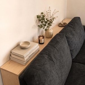Può includere: Una console in legno chiaro posizionata dietro un divano grigio scuro testurizzato. Il tavolo sostiene una pila di tre libri intitolati Kinfolk, Openhouse e Still, una piccola ciotola in ceramica, una candela accesa, un vaso con piante e un piattino.