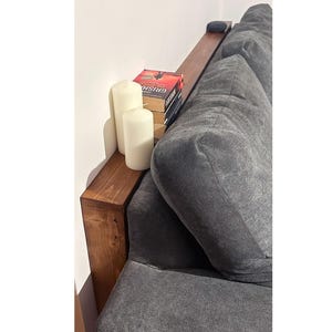 Puede incluir: Una mesa consola de madera marrón oscuro situada detrás de un sofá tapizado gris. La mesa sostiene dos velas de pilar blancas y una pila de libros, incluyendo The Appeal de John Grisham, junto con un pequeño dispositivo circular oscuro.