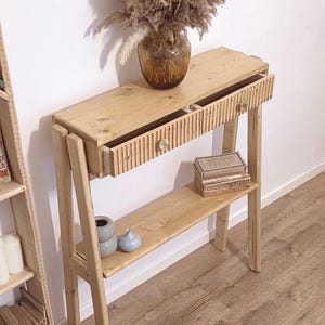 Può includere: Un tavolo consolle in legno chiaro dotato di un cassetto singolo con frontale a scanalature verticali e un piccolo pomello rotondo. Il tavolo presenta un ripiano inferiore aperto, supporti laterali inclinati e una finitura in legno naturale.