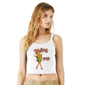 May include: White crop top with a graphic of a walking corn cob wearing red high heels. The text on the top reads "Walking -ography" in a vintage style font. The top has a scoop neckline and wide straps.