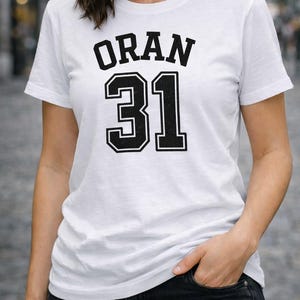 Könnte beinhalten: Weißes T-Shirt mit dem Wort "ORAN" in fetten schwarzen Buchstaben über der Nummer "31". Die Nummer ist ebenfalls schwarz mit weißem Rand. Das T-Shirt hat einen klassischen Rundhalsausschnitt.
