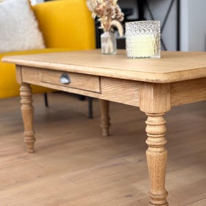 Peut inclure: Une table basse rectangulaire en chêne clair avec un seul tiroir et des pieds tournés. Un bougeoir en verre et un petit vase avec des fleurs séchées sont posés sur la table. La table est dans un salon avec un canapé jaune en arrière-plan.