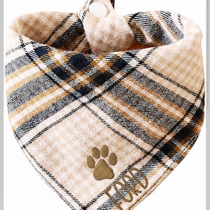 May include: A triangular pet bandana featuring a tan, black, and white plaid pattern. The fabric is embroidered with a brown paw print and the name FORD in brown thread. A white snap button is visible at the top center for fastening the accessory.
