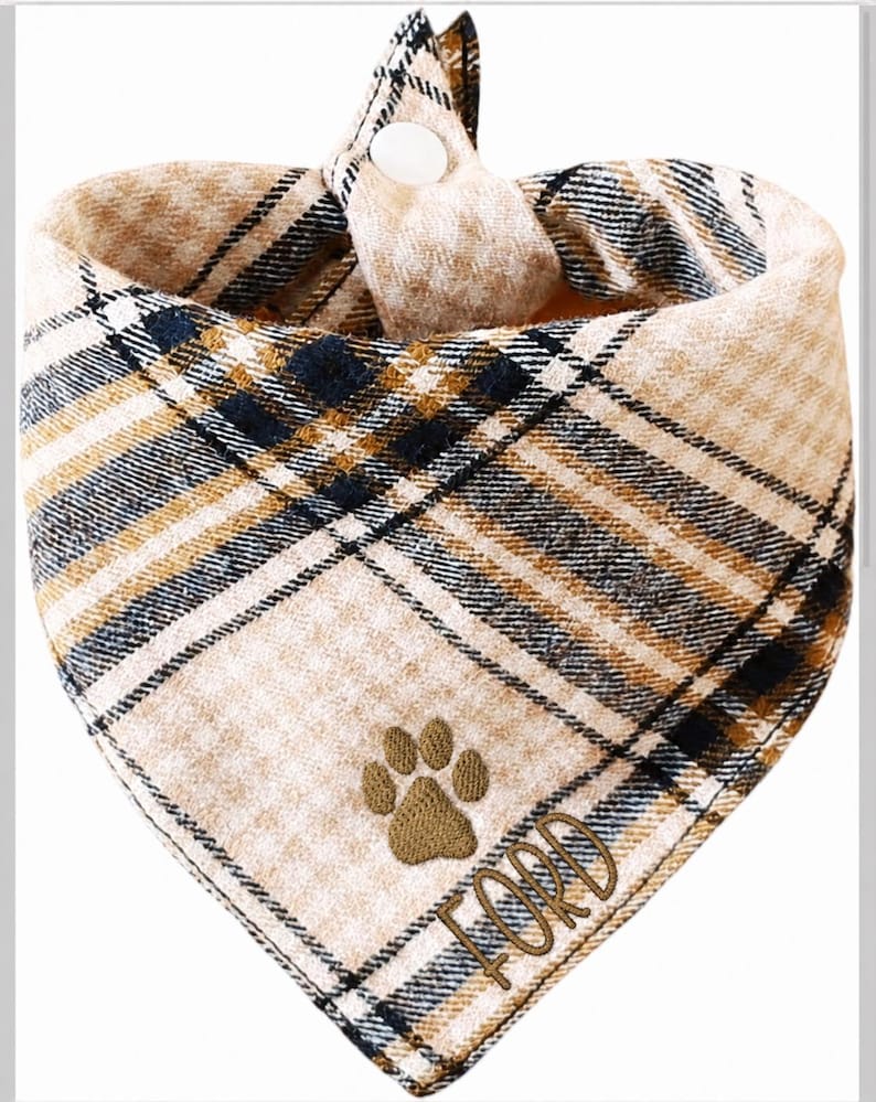 May include: A triangular pet bandana featuring a tan, black, and white plaid pattern. The fabric is embroidered with a brown paw print and the name FORD in brown thread. A white snap button is visible at the top center for fastening the accessory.