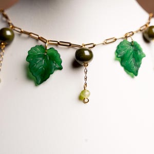 Peut inclure: Un collier à chaîne dorée orné de breloques en forme de feuilles et de perles vertes. Le collier présente des pendentifs en forme de feuilles vertes détaillées et des perles rondes, créant une esthétique inspirée de la nature. Le collier est présenté sur fond blanc.