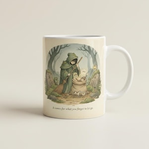 Può includere: Una tazza in ceramica bianca con l'illustrazione di una figura incappucciata in un mantello verde che tiene una scopa, vicino a un sacco pieno di piccoli oggetti e fantasmi in un bosco. Il testo recita, It comes for what you forget to let go.