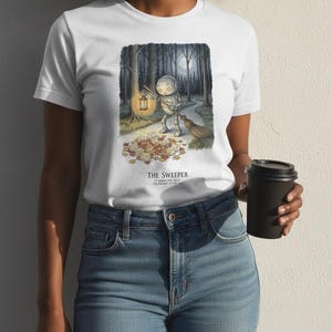 Może przedstawiać: Biały t-shirt z grafiką przedstawiającą małego robota trzymającego świecącą latarnię i miotłę, zamiatającego jesienne liście na leśnej ścieżce pod pełnią księżyca. Tekst pod ilustracją brzmi THE SWEEPER.