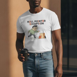 Op de afbeelding: Een wit T-shirt met korte mouwen en de tekst NULL POINTER EXCEPTION in donkergrijs en oranje. Onder de tekst staat een cartoonillustratie van een persoon met een bril in een groene hoodie die naar een zwevende doos bij andere dozen reikt.