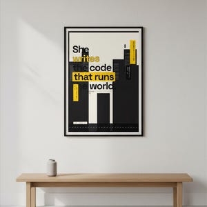 May include: A rectangular wall poster in a black frame featuring a graphic design of black city buildings against a cream background. The text reads: She writes the code that runs the world. Yellow rectangular accents highlight specific words.