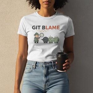 Op de afbeelding: Een wit T-shirt met korte mouwen en de tekst GIT BLAME in zwarte en oranje letters. Onder de tekst staat een afbeelding van een detective in een trenchcoat en hoed die een vergrootglas vasthoudt en naar drie kleine, ronde figuren wijst.
