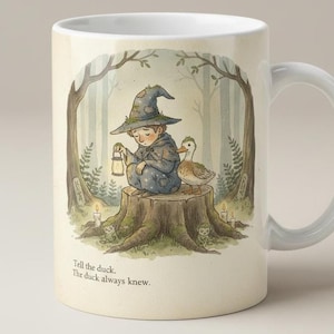 May include: A white ceramic mug featuring a printed illustration of a figure in a pointed hat sitting on a tree stump with a duck, holding a lantern. The text "Tell the duck. The duck always knew." is printed in black at the bottom of the design.