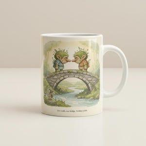 May include: A white ceramic mug featuring a printed illustration of two green trolls standing on a stone bridge over a river. The text "Two trolls, one bridge. Neither yields." is printed below the illustration. The mug has a handle on the right side.