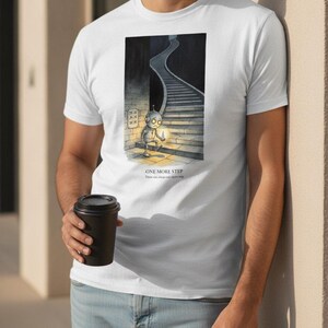 Könnte beinhalten: Ein weißes T-Shirt mit einer grafischen Illustration eines kleinen Roboters, der ein leuchtendes Licht am Fuß einer langen Treppe hält. Der Text "ONE MORE STEP" ist unter der Illustration in schwarzen Großbuchstaben mit kleinerem Text gedruckt.