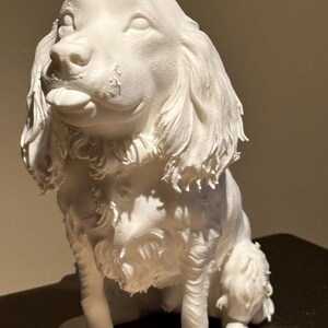 May include: A white dog sculpture, possibly a Cocker Spaniel, in a seated pose. The dog has long, flowing fur around its ears and chest, with detailed facial features and a slightly open mouth. The sculpture is made of a solid, opaque material.