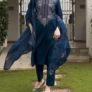 May include: A teal-colored embroidered salwar kameez ensemble. The long-sleeved tunic features intricate floral embroidery in shades of purple and green around the neckline. The outfit includes matching pants and a sheer dupatta with floral patterns. The model is wearing blue embroidered flats.