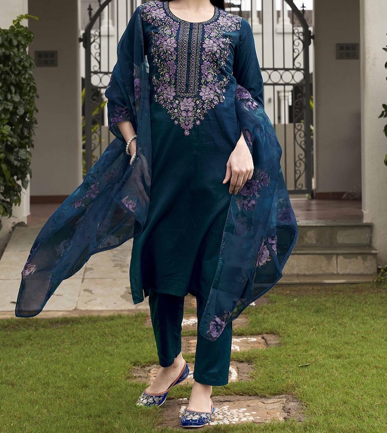 May include: A teal-colored embroidered salwar kameez ensemble. The long-sleeved tunic features intricate floral embroidery in shades of purple and green around the neckline. The outfit includes matching pants and a sheer dupatta with floral patterns. The model is wearing blue embroidered flats.