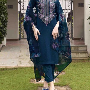 May include: A teal-colored embroidered salwar kameez suit. The long-sleeved top features intricate purple floral embroidery around the neckline and bodice. The sheer dupatta and the sleeves have a floral pattern. The suit includes matching pants and shoes.