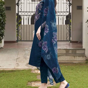 May include: A teal-colored, embroidered tunic and matching pants with a floral print shawl. The tunic features intricate purple embroidery around the neckline. The shawl has a large floral pattern in shades of purple and blue. The outfit is paired with matching teal flats.