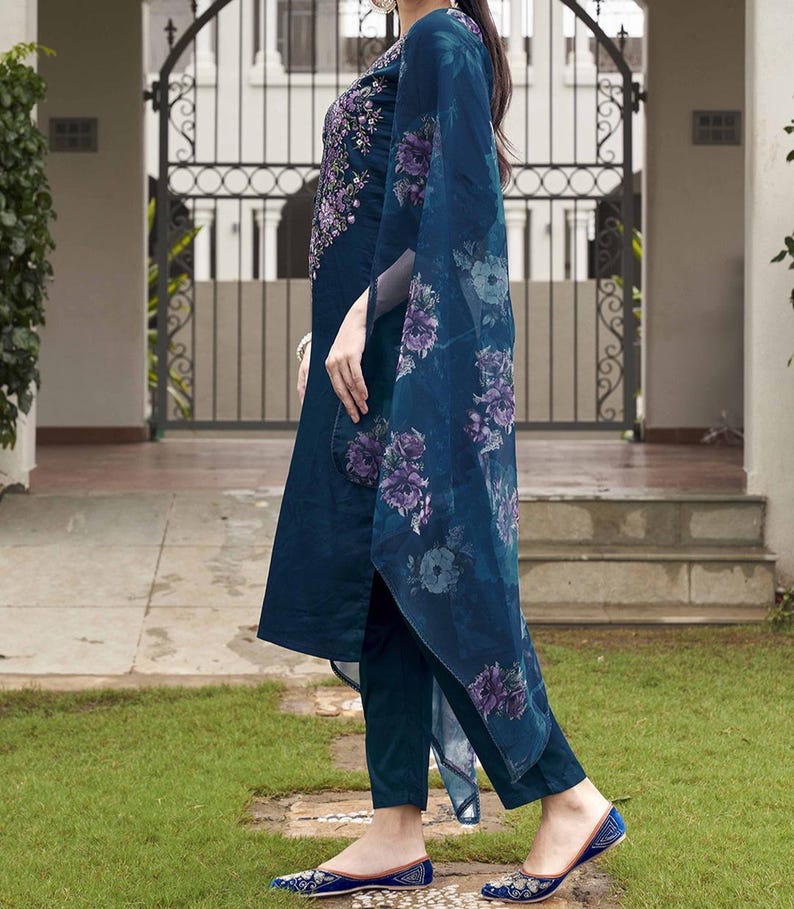 May include: A teal-colored, embroidered tunic and matching pants with a floral print shawl. The tunic features intricate purple embroidery around the neckline. The shawl has a large floral pattern in shades of purple and blue. The outfit is paired with matching teal flats.
