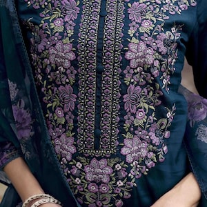 May include: A dark teal-colored tunic with intricate floral embroidery in shades of purple and green. The embroidery is concentrated around the neckline and down the center, with a buttoned placket. The sleeves are long, and a matching scarf is draped over the arm.