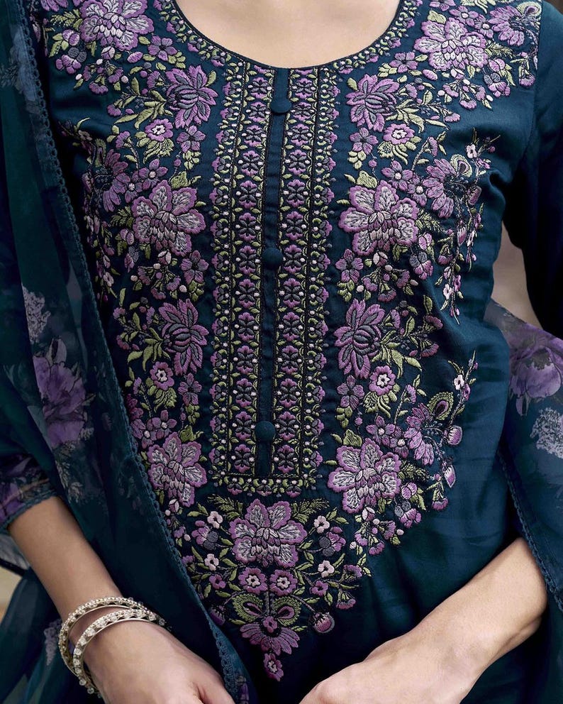 May include: A dark teal-colored tunic with intricate floral embroidery in shades of purple and green. The embroidery is concentrated around the neckline and down the center, with a buttoned placket. The sleeves are long, and a matching scarf is draped over the arm.