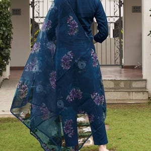 May include: A dark teal ensemble featuring a long-sleeved top, matching pants, and a sheer floral-printed scarf. The scarf and pants are adorned with purple and blue floral patterns. The outfit is worn with blue and orange sandals, set against a green lawn.