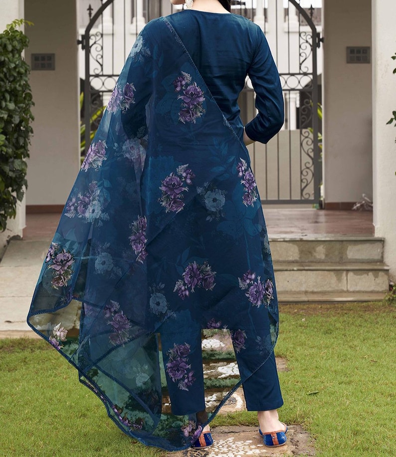May include: A dark teal ensemble featuring a long-sleeved top, matching pants, and a sheer floral-printed scarf. The scarf and pants are adorned with purple and blue floral patterns. The outfit is worn with blue and orange sandals, set against a green lawn.