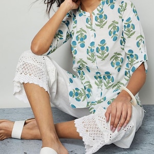 May include: A white cotton outfit with a floral print top and eyelet hem trousers. The top has a mandarin collar and button-down front. The floral pattern features blue flowers and green leaves. The trousers are cropped with a delicate eyelet design.
