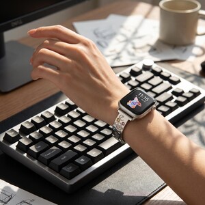 May include: A silver-toned smartwatch featuring a rectangular screen displaying a small cartoon dog graphic and the time 11:15. The watch band is metallic silver with engraved floral patterns and small colorful circular accents throughout the links.