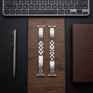 May include: A silver-toned metal watch band featuring a link design with engraved Mickey Mouse head silhouettes and small, colorful embedded crystals. The band consists of two separate segments with integrated connectors for a smartwatch attachment.
