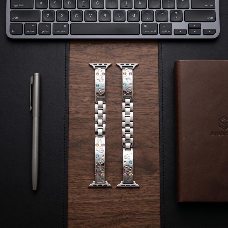 May include: A silver-toned metal watch band featuring a link design with engraved Mickey Mouse head silhouettes and small, colorful embedded crystals. The band consists of two separate segments with integrated connectors for a smartwatch attachment.