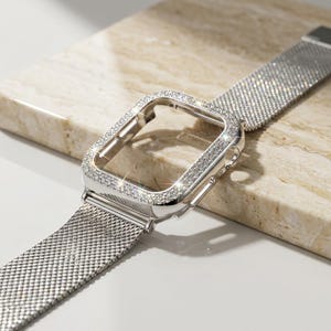 May include: A silver Apple Watch band with a matching case. The case is encrusted with small, sparkling crystals. The band is a mesh style, and the watch is displayed on a marble surface.