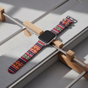 May include: A silver smartwatch with a colorful woven band featuring a geometric pattern in red, green, orange, and blue. The watch is displayed on a gray surface with wooden supports.