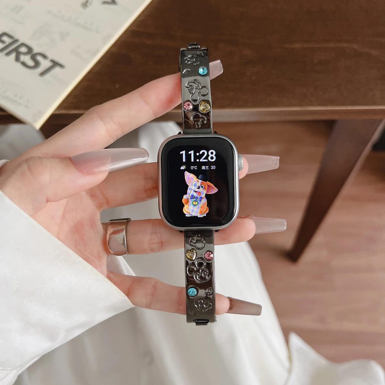 May include: A dark metallic smartwatch band featuring embossed mouse head silhouettes and embedded multicolored crystals. The watch face displays the time 11:28 and a colorful illustration of a dog wearing a hat and bowtie against a black background.