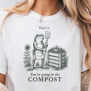 May include: A white short-sleeve t-shirt featuring a dark green graphic of a frog wearing an apron and holding a pitchfork next to a compost bin and sunflower. The text reads: That's it. You're going in the COMPOST. The shirt is paired with denim shorts.