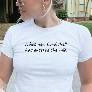 May include: A white short-sleeved t-shirt featuring the black printed text 'a hot new bombshell has entered the villa' centered on the chest. The shirt is paired with light blue denim jeans, a gold-toned bracelet, and gold-toned rings.