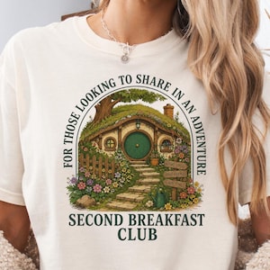 May include: An off-white short-sleeve t-shirt featuring a graphic of a round-door cottage nestled in a grassy hill. The text reads: FOR THOSE LOOKING TO SHARE IN AN ADVENTURE SECOND BREAKFAST CLUB. The design includes flowers, a fence, and a stone path.
