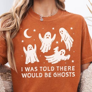 May include: A burnt orange short-sleeve t-shirt featuring a white graphic print of four ghosts, stars, and a crescent moon. The text "I WAS TOLD THERE WOULD BE GHOSTS" is printed in white capital letters below the ghost illustrations.